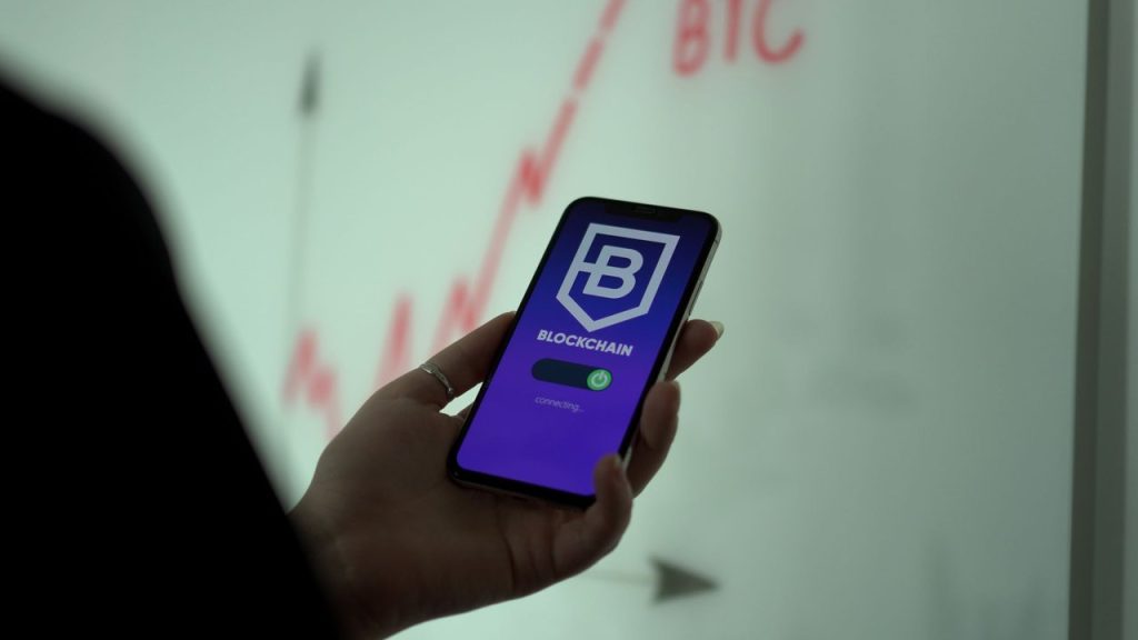 Mão segurando um smartphone com um aplicativo de blockchain e gráfico financeiro exibido na tela.