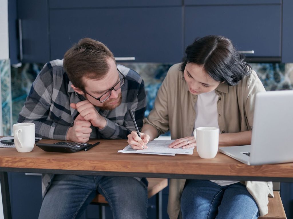 Quando a dívida compromete o básico: alerta do Banco Central sobre as famílias brasileiras Casal analisa documentos financeiros com um laptop e uma calculadora à mesa da cozinha.