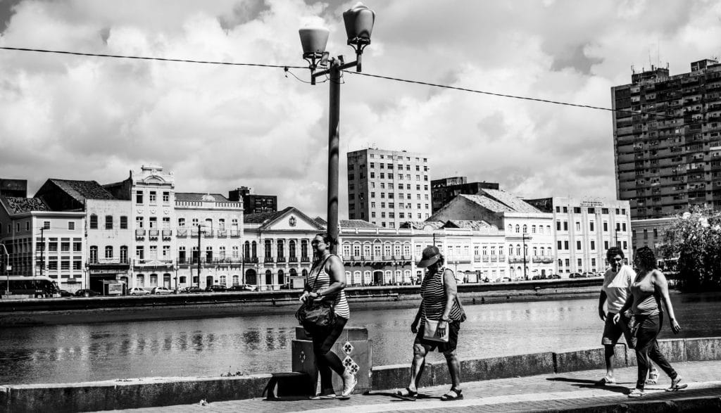Recife testa tecnologia que transforma caminhadas e pedaladas em recompensas Pessoas caminham diante de prédios históricos do Recife em uma cena urbana em preto e branco.